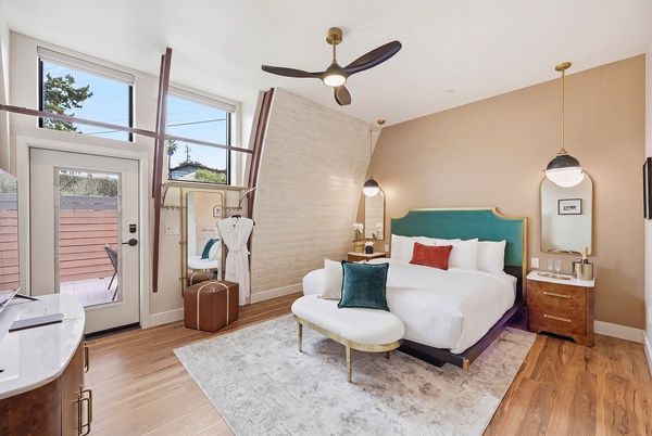 Bright, modern bedroom with a slanted ceiling, large windows, and a glass door leading to a patio. The room features a king-sized bed with a teal upholstered headboard, white bedding, and accent pillows. A curved bench sits at the foot of the bed on a light area rug. Warm wood floors, a ceiling fan, pendant lights, and mid-century style furniture complete the airy, stylish space.