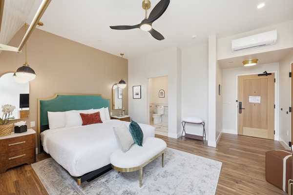 Bright, modern bedroom with a slanted ceiling, large windows, and a glass door leading to a patio. The room features a king-sized bed with a teal upholstered headboard, white bedding, and accent pillows. A curved bench sits at the foot of the bed on a light area rug. Warm wood floors, a ceiling fan, pendant lights, and mid-century style furniture complete the airy, stylish space.
