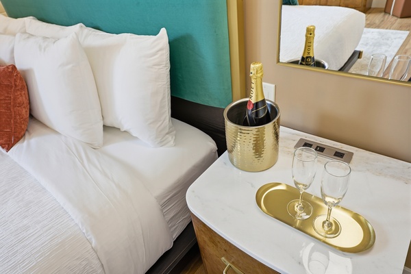 Close-up of a king-sized bed with a teal upholstered headboard and white bedding, next to a bedside table featuring a gold ice bucket with a bottle of champagne, two champagne flutes on a gold tray, and a wall mirror reflecting the setup.