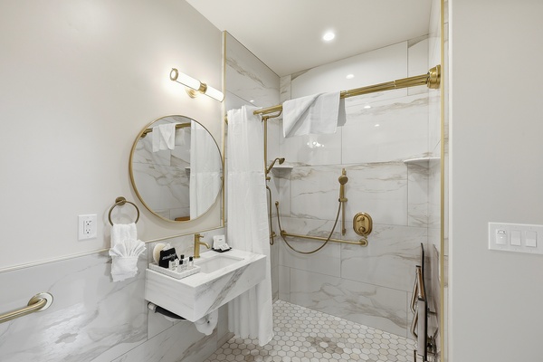 Modern ADA compliant bathroom with a walk-in glass shower featuring white marble-style tile walls and gold fixtures, including a handheld showerhead and grab bar. A floating white sink with a gold faucet sits beneath a round mirror, with neatly arranged toiletries and folded white towels. The space is finished with hexagonal tile flooring and soft, warm lighting for a clean, upscale look.