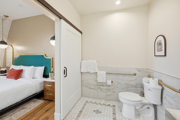 Accessible bathroom with a white toilet, gold grab bars, and marble-style wall tile, featuring hexagonal tile flooring and neatly hung white towels. A sliding door opens to an adjacent bedroom with a king-sized bed, teal upholstered headboard, and warm wood flooring, creating a cohesive and modern suite layout.