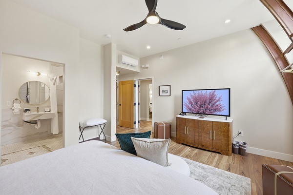 Modern connecting hotel room with a king-sized bed in the foreground, featuring white bedding and accent pillows, facing a wooden dresser with a flat-screen TV. The space includes warm wood flooring, a ceiling fan, and neutral walls. An open doorway reveals a bathroom with a floating sink and round mirror, while a connecting door leads to an adjoining room, creating a bright and spacious suite layout.