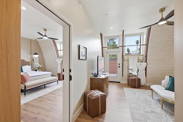 Modern connecting hotel rooms with an open doorway between them, both featuring king-sized beds with white bedding and accent pillows. The spaces include warm wood flooring, vaulted ceilings, and contemporary furnishings, with bench seating, large windows, ceiling fans, and a glass door leading to private patios, creating a bright and cohesive dual-room layout.