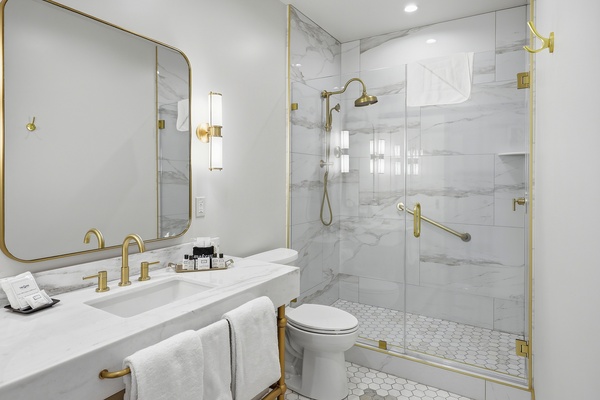 Bright, modern hotel bathroom with a glass-enclosed walk-in shower featuring marble-style tile, a handheld and rainfall showerhead, and a built-in grab bar. The space includes a white vanity with an integrated sink, gold fixtures, a large rounded-edge mirror, and wall sconces. A toilet, hex tile flooring, and neatly arranged towels and amenities complete the clean, upscale design.