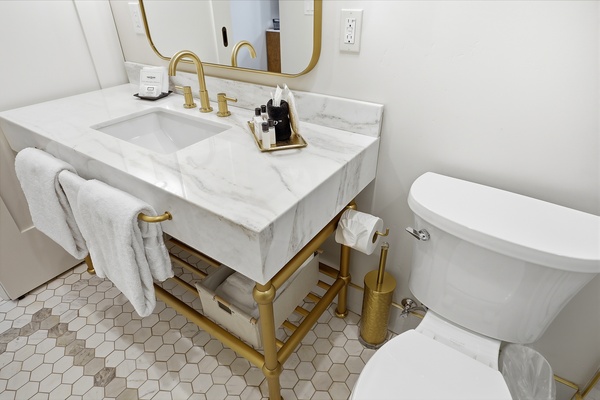 Modern hotel bathroom vanity with a white marble countertop and integrated sink, complemented by gold fixtures and a large rounded mirror. The space includes neatly folded towels, upscale bath amenities, and a hex tile floor, with a toilet positioned next to the vanity for a clean, compact layout.