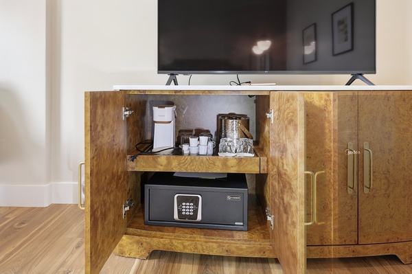 Wood cabinet with open doors beneath a flat-screen TV, revealing a coffee station with a single-serve coffee maker, cups, and glassware, alongside an ice bucket. A secure in-room safe is stored on the lower shelf, with warm wood flooring and brass hardware completing the modern hotel room setup.