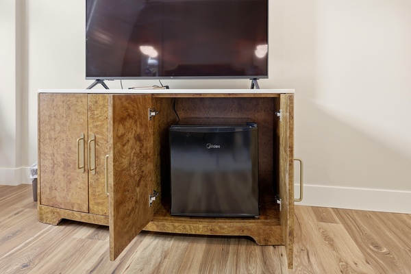 Wood cabinet beneath a flat-screen TV with one door open, revealing a compact mini refrigerator inside. The cabinet features a warm burl wood finish with brass handles, set against light walls and wood flooring in a modern hotel room.