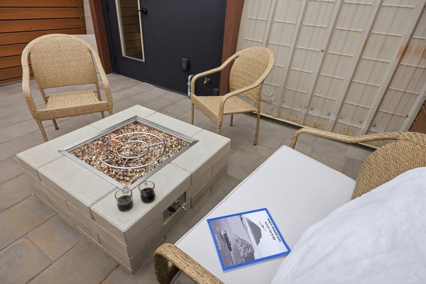 Private outdoor patio with a square fire pit table at the center, surrounded by woven wicker chairs with light cushions. Two glasses of red wine sit beside the fire, and a book rests on a cushioned lounge chair, creating a cozy and intimate seating area with modern finishes.