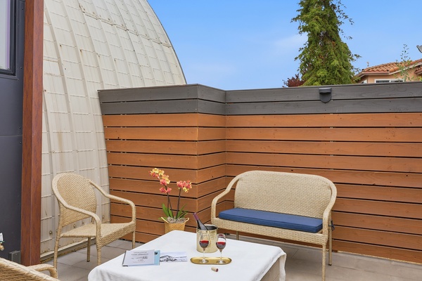 Private outdoor patio seating area with woven wicker chairs and a small sofa arranged around a cushioned table. A bottle of wine in an ice bucket and two glasses sit on a tray alongside a book, while a potted orchid adds a decorative touch. The space is enclosed by wood-paneled fencing with a curved architectural wall, creating a cozy and stylish outdoor retreat.