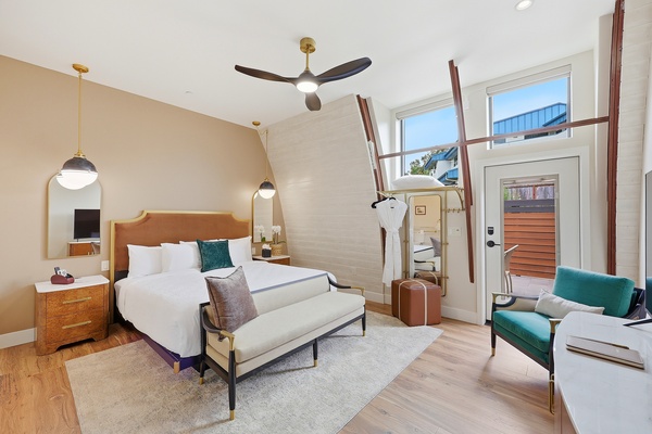 Bright, modern hotel room with a king bed featuring a teal upholstered headboard and matching bench, flanked by wood nightstands and globe pendant lights. The space includes a high ceiling with a contemporary ceiling fan, large windows, and a glass door leading to a private patio with outdoor seating. A luggage rack with robe, accent chair, and neutral rug complete the airy, stylish design.