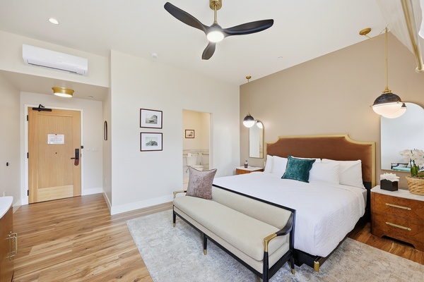 Modern hotel room with a king bed featuring a warm tan upholstered headboard, white bedding, and accent pillows, plus a cushioned bench at the foot. The space includes wood flooring, a neutral area rug, mid-century style nightstands, and globe pendant lighting. A ceiling fan and wall-mounted air unit add comfort, while the entry door and minimalist wall art complete the clean, contemporary layout.