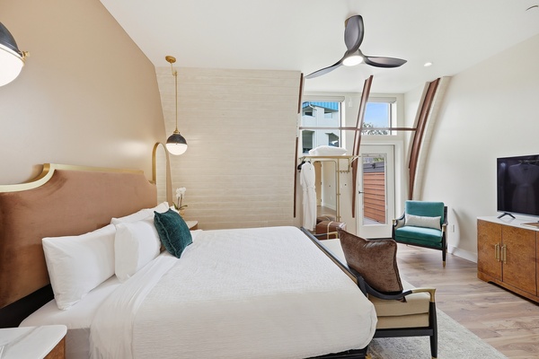 Bright, modern bedroom with a slanted ceiling, large windows, and a glass door leading to a patio. The room features a king-sized bed with a teal upholstered headboard, white bedding, and accent pillows. A curved bench sits at the foot of the bed on a light area rug. Warm wood floors, a ceiling fan, pendant lights, and mid-century style furniture complete the airy, stylish space.