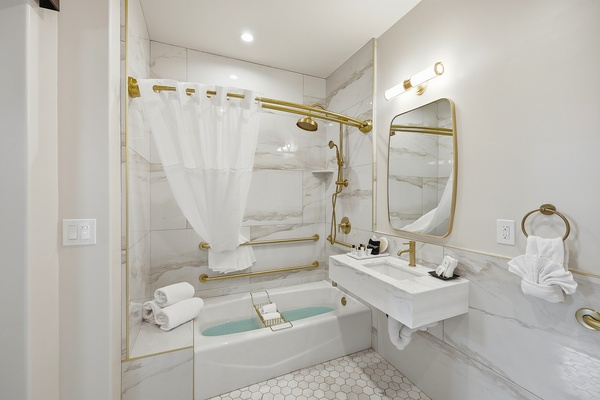ADA-compliant bathroom with a combined bathtub and shower, featuring white marble-style tile walls, gold fixtures, and multiple grab bars for accessibility. The space includes a floating sink with a gold faucet beneath a rounded mirror and wall-mounted light, along with neatly arranged towels and toiletries, all designed for a clean, modern, and accessible layout.
