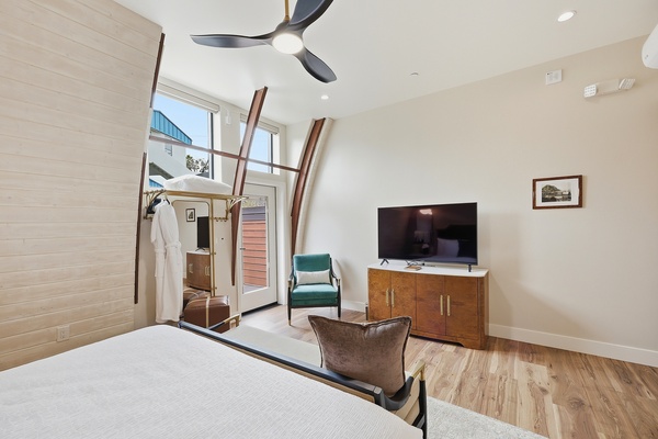 Bright, modern bedroom with a slanted ceiling, large windows, and a glass door leading to a patio. The room features a king-sized bed with a teal upholstered headboard, white bedding, and accent pillows. A curved bench sits at the foot of the bed on a light area rug. Warm wood floors, a ceiling fan, pendant lights, and mid-century style furniture complete the airy, stylish space.