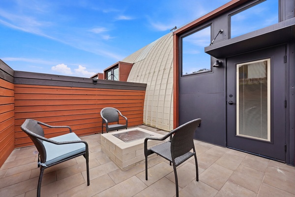 Private outdoor patio with modern design, featuring three woven chairs arranged around a square fire pit table on tiled flooring. The space is enclosed by wood-paneled fencing and includes a glass door leading inside, with a curved architectural roofline and blue sky visible above, creating a clean and contemporary outdoor seating area.